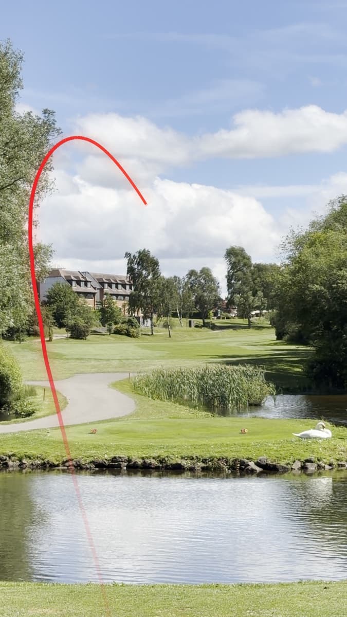 A tee shot over water on a parkland hole with a red shot tracer line rising from the tee and curving down to the left